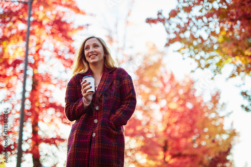 young blond woman with coffee standing under red autumn trees smiling