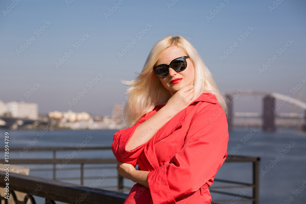 Fashionable blonde model posing in red cloak, wears sunglasses on a blurred bridge background Fashionable blonde model posing in red cloak, wears sunglasses on a blurred bridge background