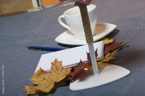 Forged metal candlestick with candles. There is an open notepad and a pen. A cup with unapproved coffee. Fallen autumn leaves of yellow and red are scattered on the surface. photo