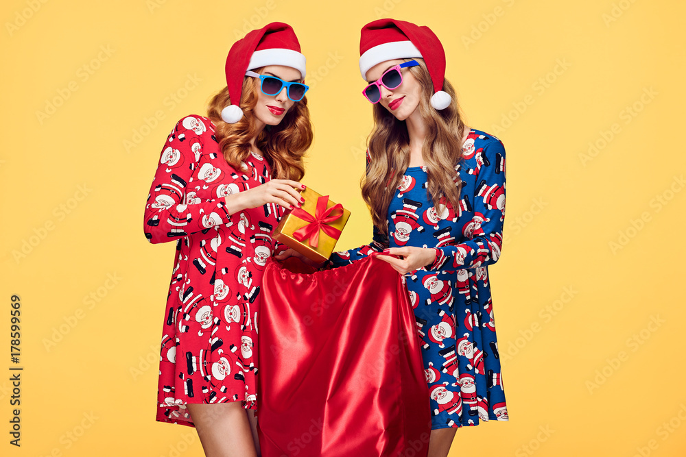 Christmas. Young Woman and Santa Sack with Presents Having Fun Happy Smiling. New Year Fashion. Best Friends Twins in Stylish fashion Red Xmas Holiday Dress on Yellow.Christmas Colorful
