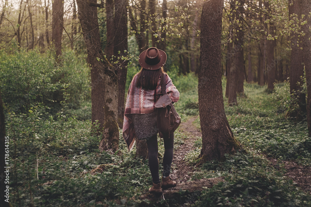 stylish hipster traveler girl in hat with backpack walking in woods in evening sunshine. bohemian fashionable woman exploring in sunlight. space for text. atmospheric moment. wanderlust