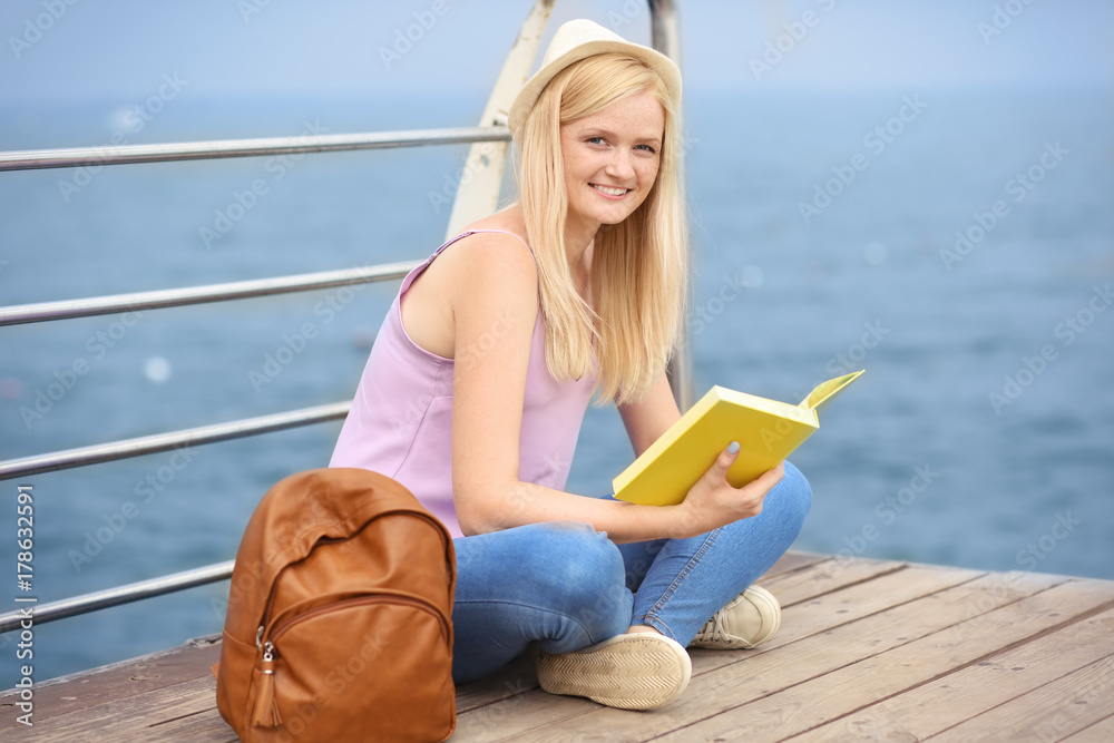 Beautiful young woman reading book while sitting on pier