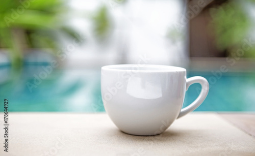 White ceramic coffee cup over blurred swimming pool, outdoor day light