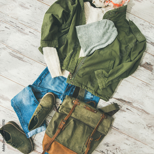 Flat-lay of Fall lady' s clothes outfit. Ecru colored knitted woolen sweater, parka, linen scarf, denim jeans, haki suede boots, backpack, cap over parquet background, top view, square crop photo