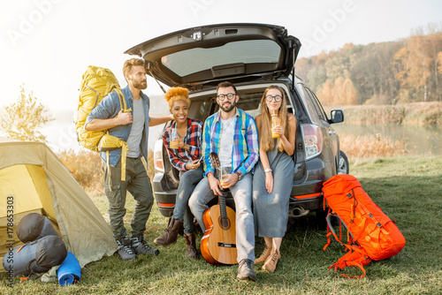 Multi ethnic group of friends sitting at the car trank during the outdoor recreation with tent and hiking equipment near the forest photo