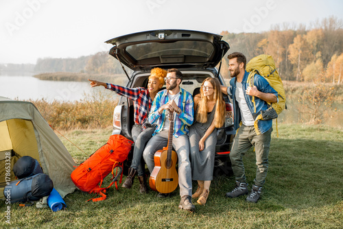 Multi ethnic group of friends sitting at the car trank during the outdoor recreation with tent and hiking equipment near the forest photo