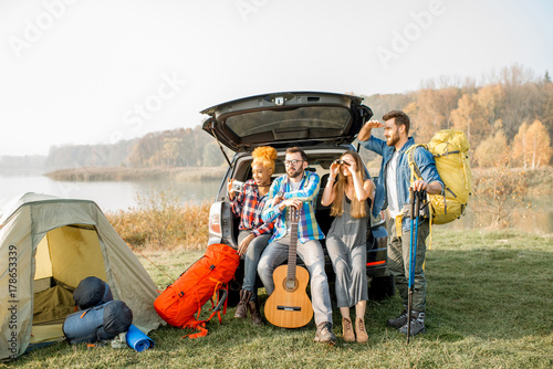 Multi ethnic group of friends sitting at the car trank during the outdoor recreation with tent and hiking equipment near the forest photo