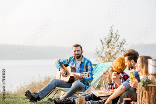 Multi ethnic group of friends dressed casually having a picnic, playing guitar and eating pizza, during the outdoor recreation near the lake © rh2010