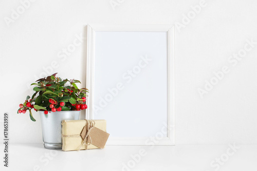 White blank wooden frame mockup with potted red teaberry plant and package with a gift tag on the white table. Styled stock feminine photography. Home decor. Christmas winter concept.
