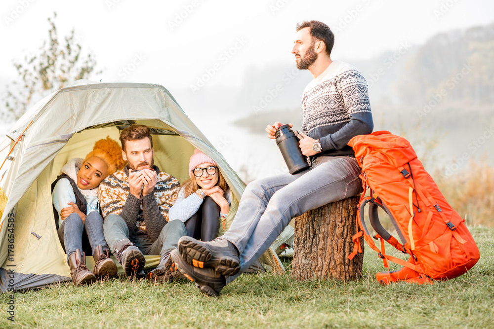 Multi ethnic group of friends dressed in sweaters warming up together sitting during the outdoor recreation near the tent Multi ethnic group of friends dressed in sweaters warming up together sitting during the outdoor recreation near the tent