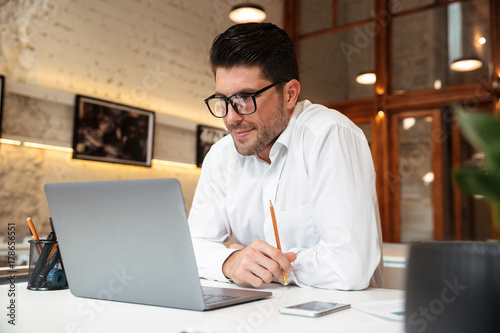 Cheerful unshaven businessman in white shirt looking at laptop screen in office