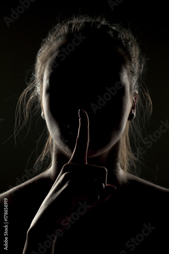 Misterious portrait of woman in shadow with finger on her lips on dark background photo
