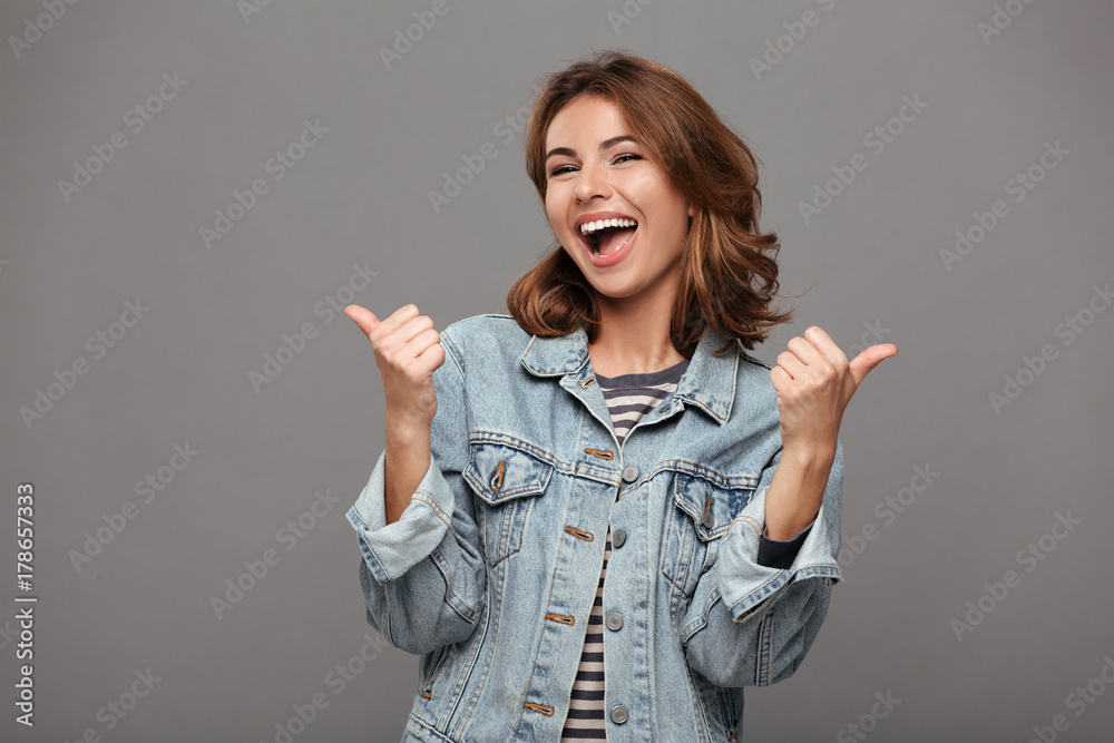 Happy attractive brunette woman in jeans jacket showing thumb up gesture with two hands, looking at camera