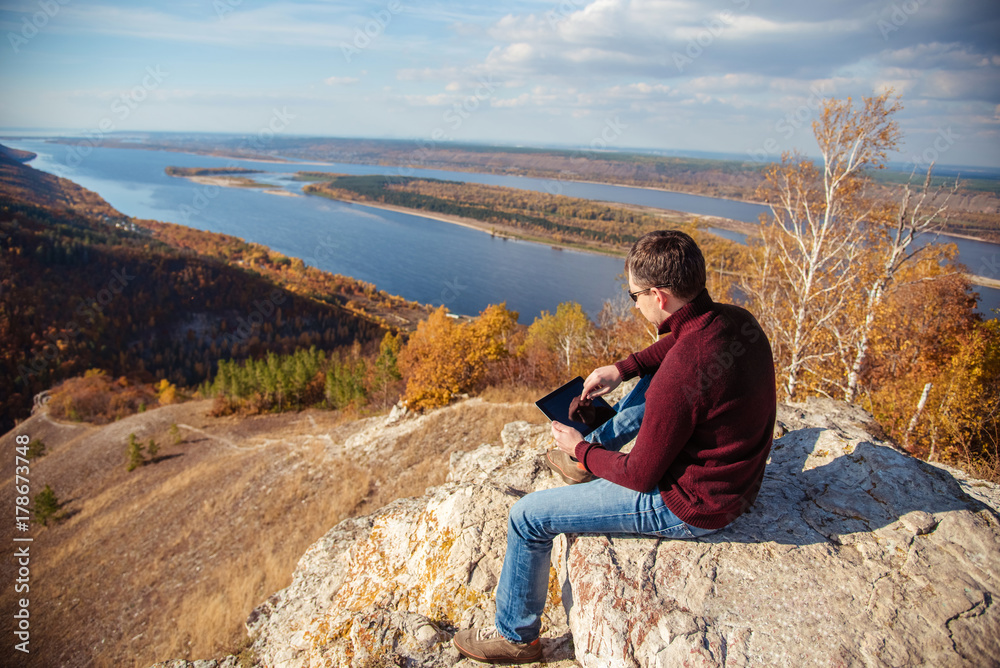 a man sits on top of a mountain, admires the scenery and works on a tablet