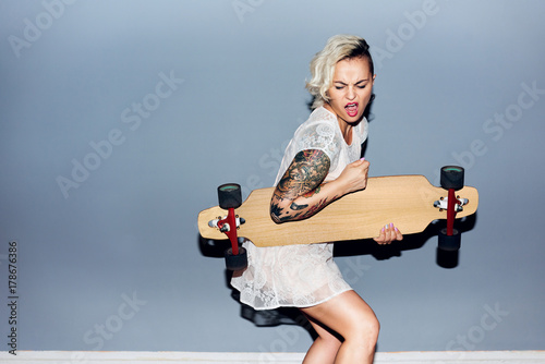 Tattooed pretty blond girl goofing around, playing longboard as guitar and singing photo