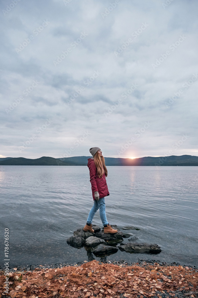 Hipster woman at the lake shore looking at beautiful sunset over the mountains Hipster woman at the lake shore looking at beautiful sunset over the mountains