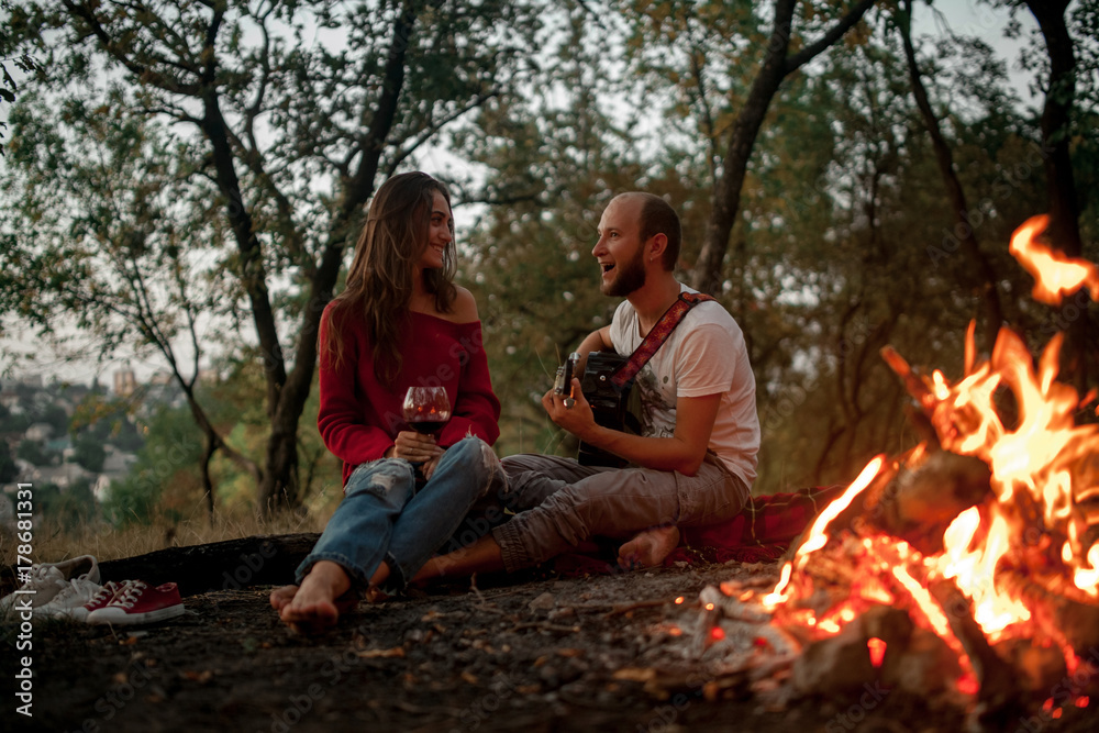 Young man plays on guitar and sings song near girl against background of bonfire flame. Young man plays on guitar and sings song near girl against background of bonfire flame.