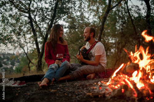 Young man plays on guitar and sings song near girl against background of bonfire flame.