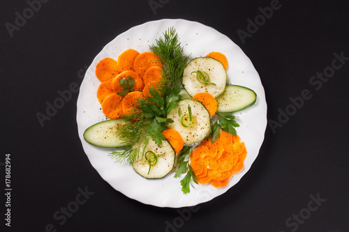 Festive cutting from fresh vegetables with greens and black pepper on a white plate on a black background.