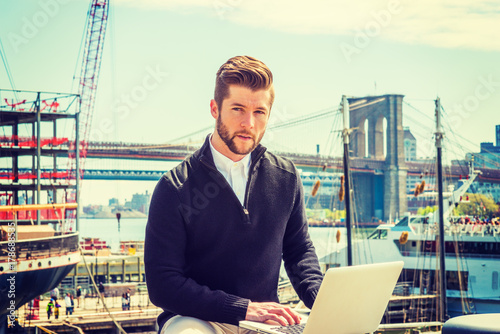 Obraz na płótnie American businessman with beard traveling, working in New York, wearing black sweater, tan pants, sitting on bench at deck on harbor, working on laptop computer. Brooklyn bridge, boat on background.. w salonie