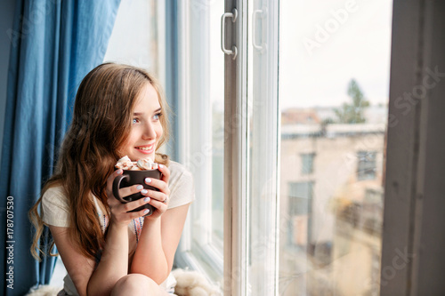 Beautiful young woman drinking coffee and looking through window while sitting at windowsill at home