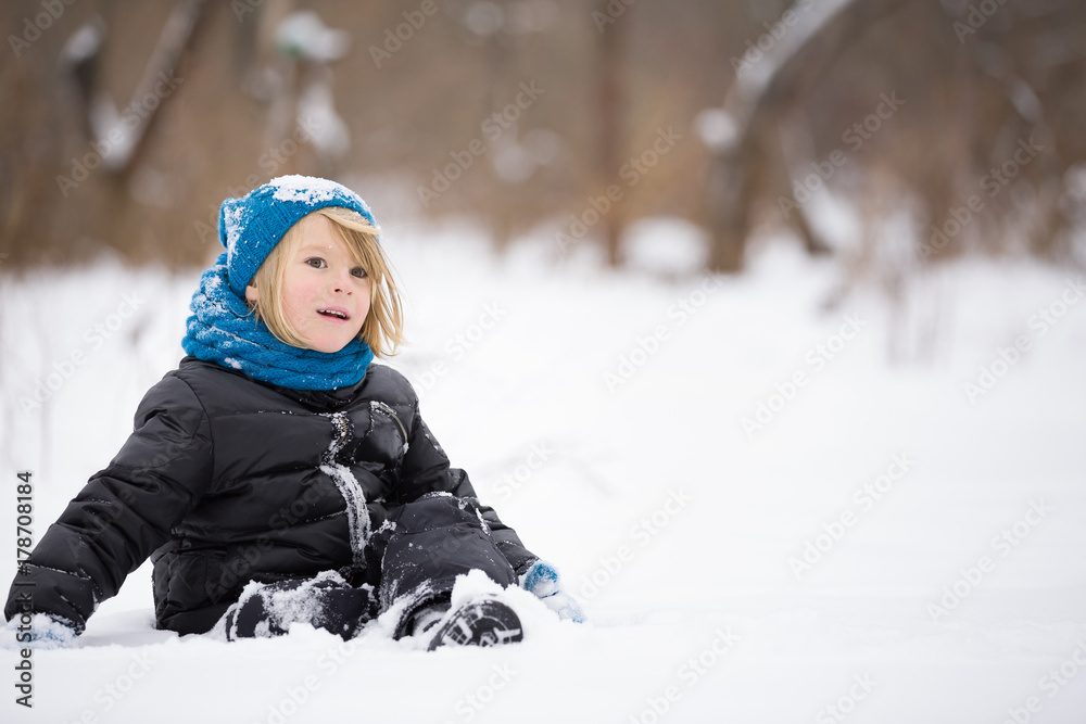 Portrait of cute kid boy in warm clothes and hat sitting on the snow and playing with it in the park. Child having fun outdoors. Winter, lifestyle concept.