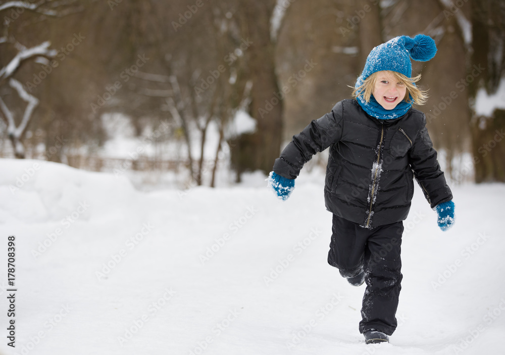 Portrait of adorable little kid boy with long blond hair running and playing with snow outdoors in the park. Child with blue scarf and hat walking and having fun on a windy winter day. Portrait of adorable little kid boy with long blond hair running and playing with snow outdoors in the park. Child with blue scarf and hat walking and having fun on a windy winter day.