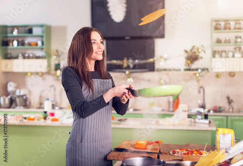 Attractive young brunette housewife in casual clothes and apron tossing pancakes on pan in the kitchen