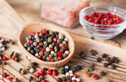 Mix of peppercorns in wooden spoon on rustic background with pink salt in glass jar. Red, black, green and white dry pepper in seeds.