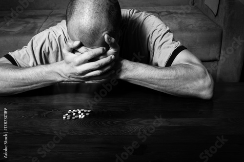 Depressed man suffering from suicidal depression want to commit suicide by taking strong medicament drugs and pills while he is sitting on the floor of his dark room black and white image