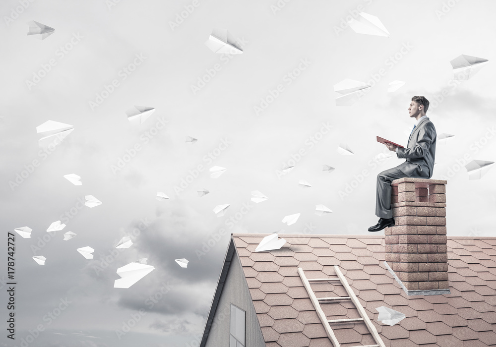 Man on brick roof reading book and paper planes flying in air Man on brick roof reading book and paper planes flying in air