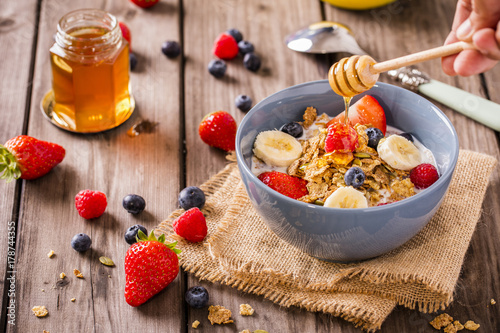 breakfast cereal shot on linen cloth wood boards angled view with raspberries blueberries sliced bananas strawberries with hand swirling honey landscape