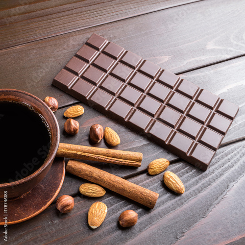 Hot black coffee in brown clay cup, dark chocolate bar, almond, hazelnut and cinnamon stick on wooden background