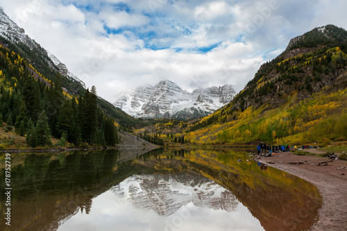 Maroon Peak and its reflection on Maroon Lake and aspen trees with its gold yellow leaves in fall foliage autumn season in a bright day light sunny day cloudy blue sky, Aspen, Colorado, USA.