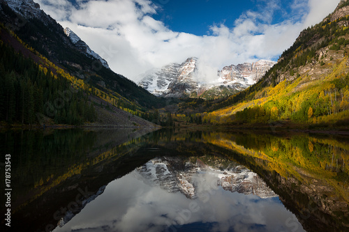 Maroon Peak and its reflection on Maroon Lake and aspen trees with its gold yellow leaves in fall foliage autumn season in a bright day light sunny day cloudy blue sky, Aspen, Colorado, USA.