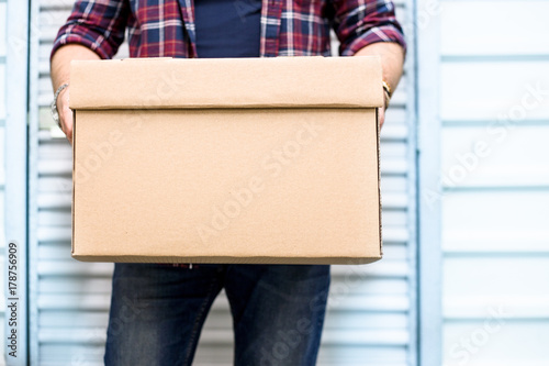 Young man holding a moving cardboard box in front of a storage door.Life style, storage, moving, storing, organizing concept. Space to write photo