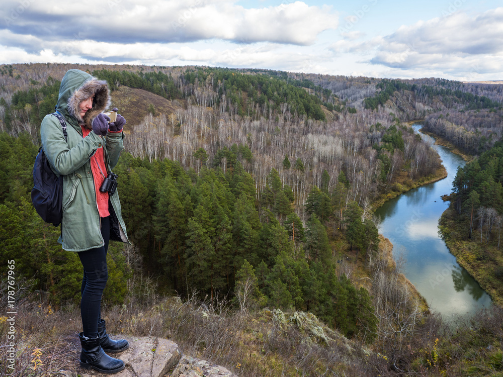 A girl on the edge of a mountain ledge photographs a beautiful turn of the river. Camp style, binoculars and backpack.