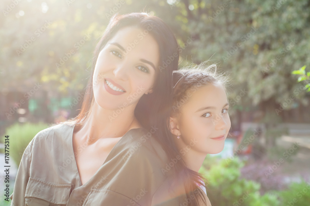 Young mom and daughter sitting back to back in park on sunny day Young mom and daughter sitting back to back in park on sunny day