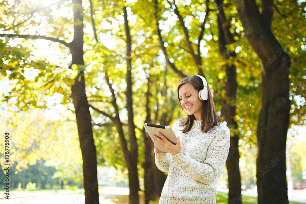 A beautiful happy cheerful brown-haired woman in white sweater with a tablet listening music in the white headphones in fall park on a warm day. Autumn in the city. A beautiful happy cheerful brown-haired woman in white sweater with a tablet listening music in the white headphones in fall park on a warm day. Autumn in the city.