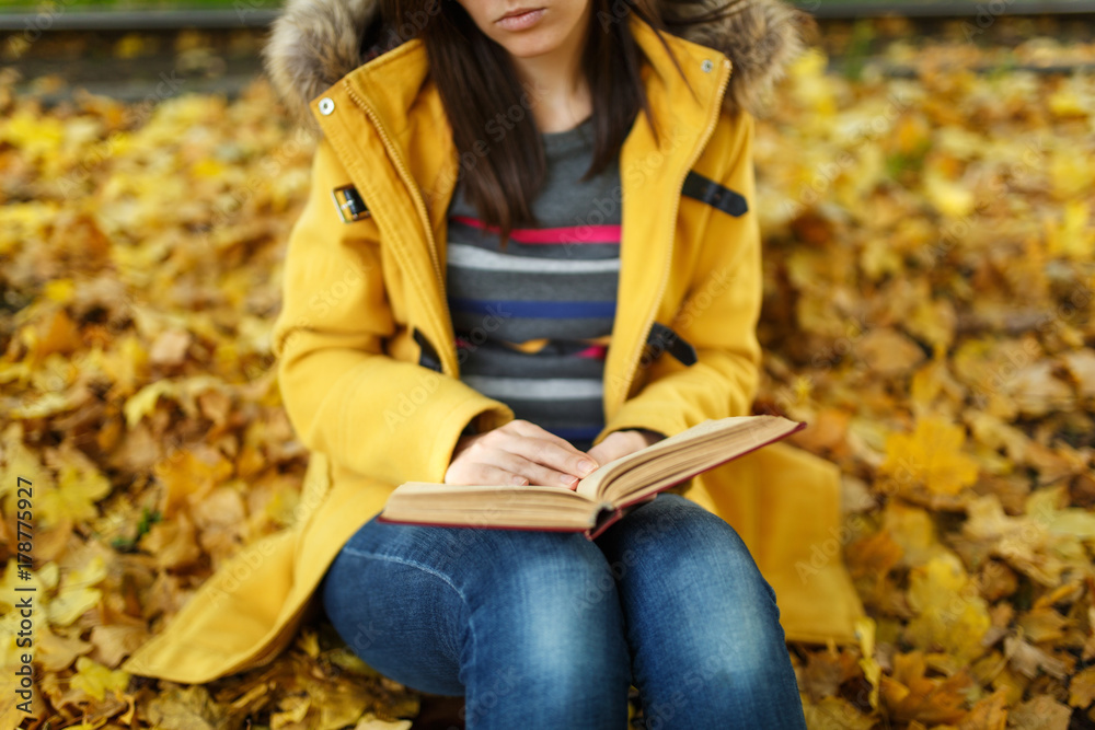 Obraz premium Woman in yellow coat and jeans sitting under the maple tree with a red book in fall city park on a warm day. Autumn golden leaves. Reading concept. Close up.