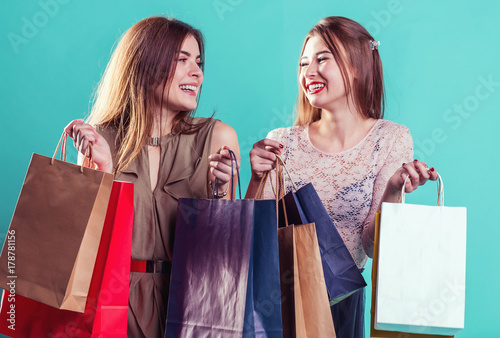 Friends girls smiling holding colorful shopping bags before blue indoor background, shopaholic concept