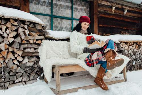 Woman enjoying time on her own on a cold winter day. Winter relax - woman sitting outside a cottage and reading a book on a snowy day.