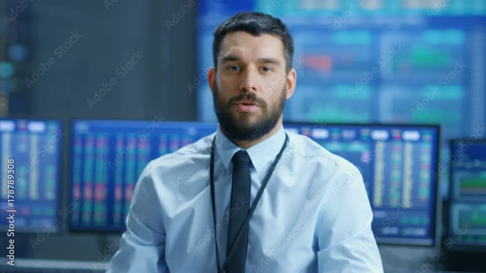 Professional Stock Market Trader Charismatically Talks into the Camera. Behind Him Computer Screens with Ticker Numbers, Data, Graphs. Shot on RED EPIC-W 8K Helium Cinema Camera.