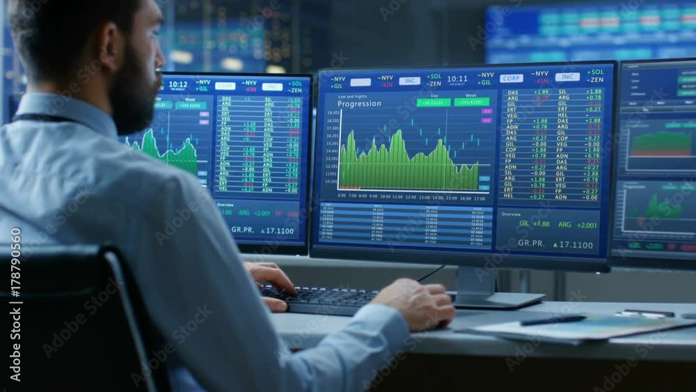 Over the Shoulder View of Stock Market Trader Working on a Computer with Multiple Monitors Showing Stock Ticker Numbers and Graphs. Shot on RED EPIC-W 8K Helium Cinema Camera. Over the Shoulder View of Stock Market Trader Working on a Computer with Multiple Monitors Showing Stock Ticker Numbers and Graphs. Shot on RED EPIC-W 8K Helium Cinema Camera.