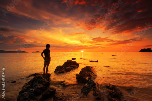 silhouette of young boy standing on rock in the sea with sunset sky, long speed exposure,Tarn-khu beach at Trat, Thailand