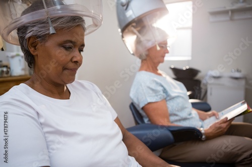 Senior women reading magazine while sitting under hair steamer