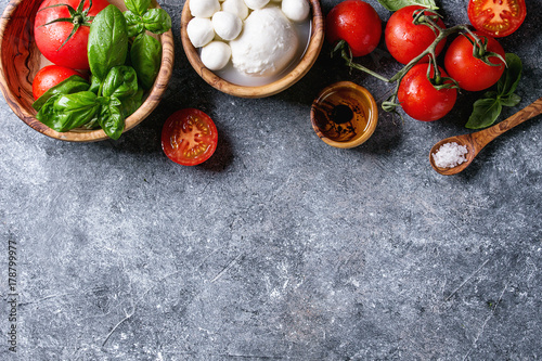Ingredients for italian caprese salad. Mozzarella balls, buffalo, tomatoes, basil leaves, olive oil with vinegar, salt in olive wood bowls over gray texture background. Top view with copy space
