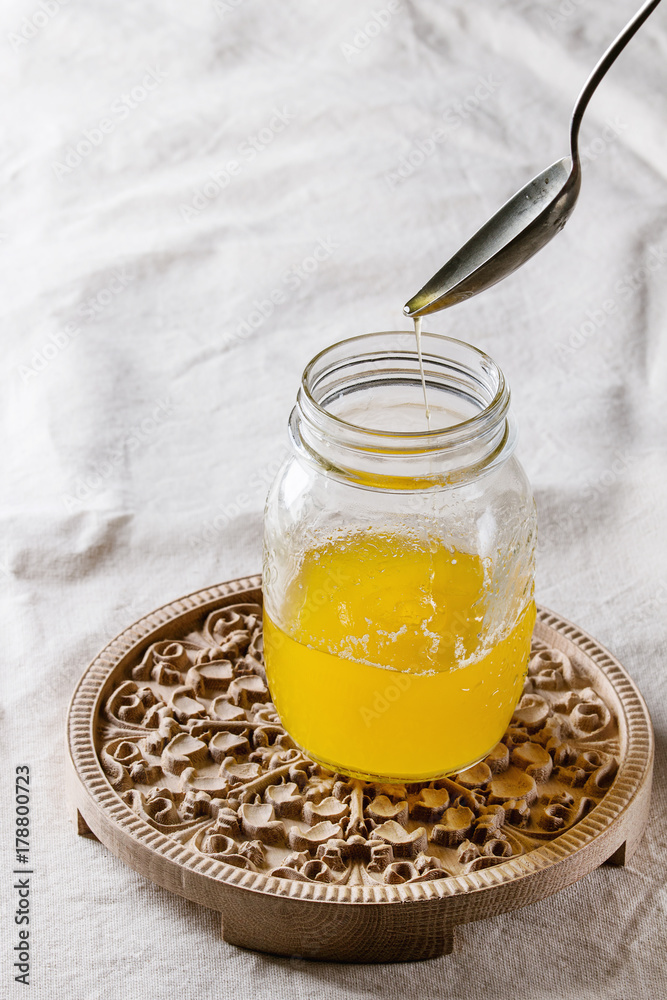 Glass of hot drawn ghee butter standing on wooden carved ornate board with pouring butter from spoon over white linen textile tablecloth. Healthy eating
