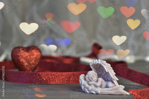 Fototapeta Naklejka Na Ścianę i Meble -  A figurine of a small sleeping angel. Background red heart and hearts-bokeh. St. Valentine's Day