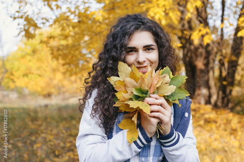 Girl standing in autumn park and holding in hands bunch of leaves. Young woman looking at camera and smile. Outdoor. Close up. Autumn.